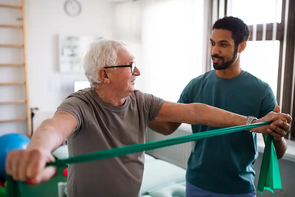 An older man uses a green resistance band for arm exercises while a younger man assists him in a brightly lit room.