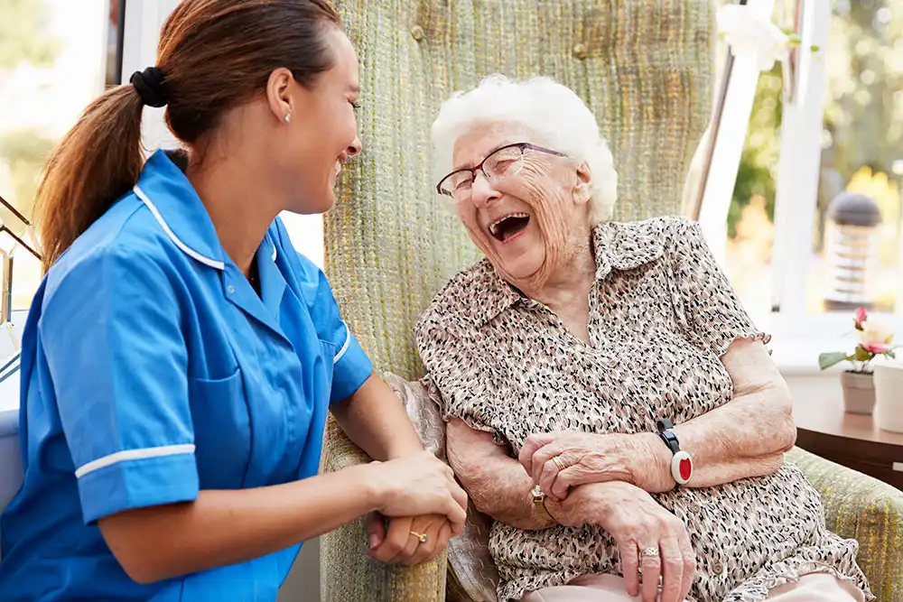 A smiling caregiver in a blue uniform sits beside an elderly woman in a chair; both are laughing and holding hands in a bright indoor setting. A smiling caregiver in a blue uniform sits beside an elderly woman in a chair; both are laughing and holding hands in a bright indoor setting.