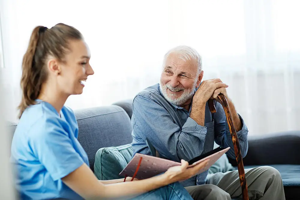 A young caregiver sits on a couch with an elderly man holding a cane, showing him a book or folder as they both smile.