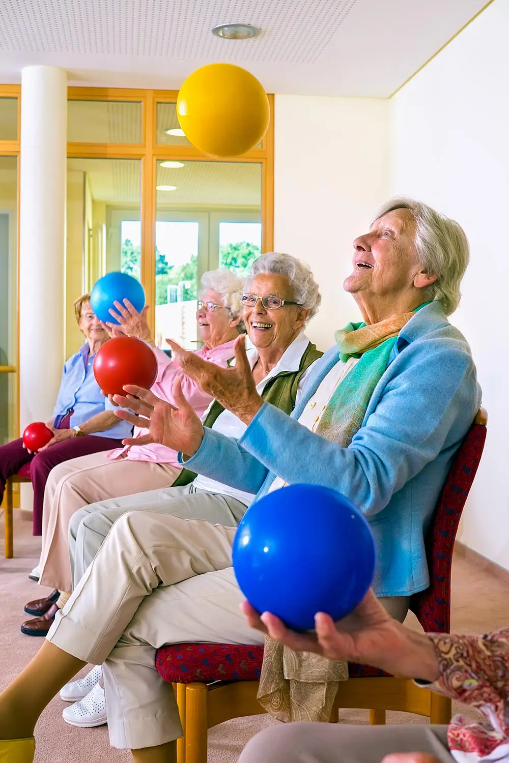 A group of elderly women sitting indoors, tossing colorful balls into the air as part of a seated exercise or activity session.