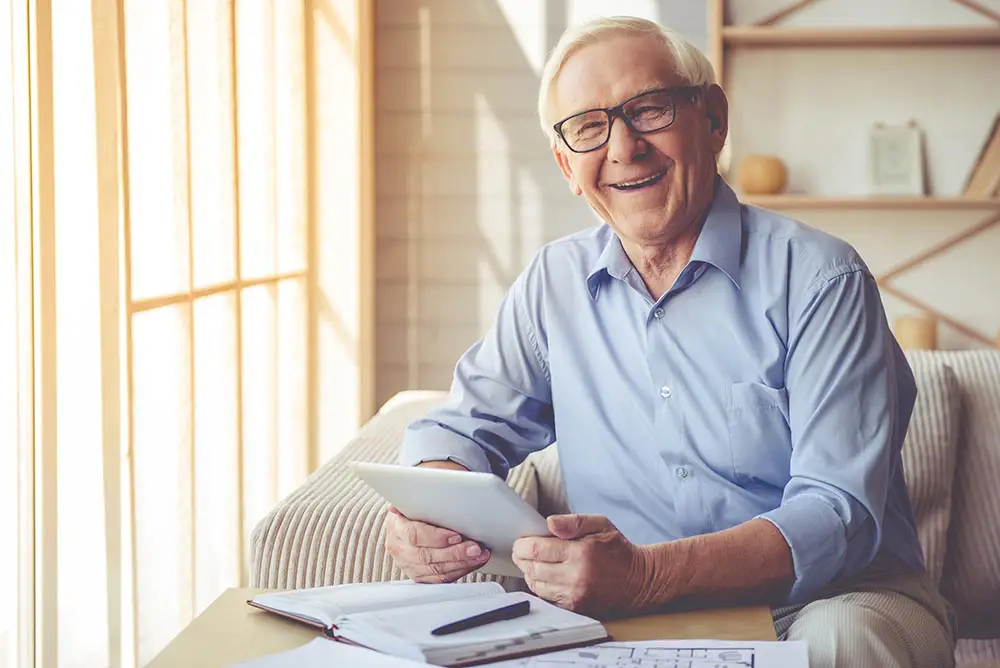 Older man with glasses sits on a couch, smiling, holding a tablet, with papers, pen, and eyeglasses on the table in front of him near a bright window.