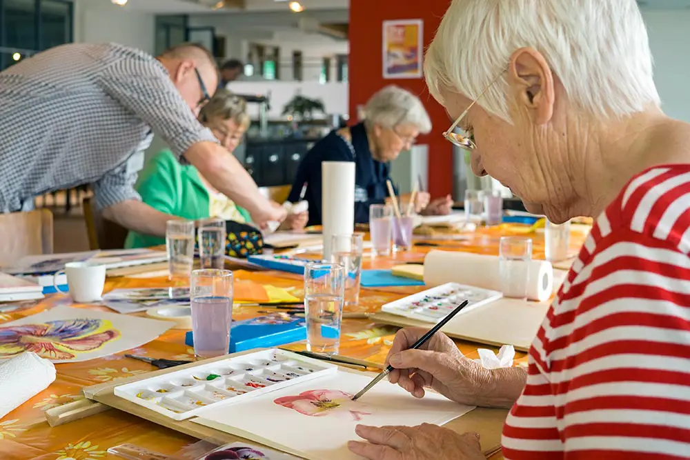 Several seniors sit at a table engaged in watercolor painting, with art supplies and glasses of water spread out on the table in a bright, communal space.