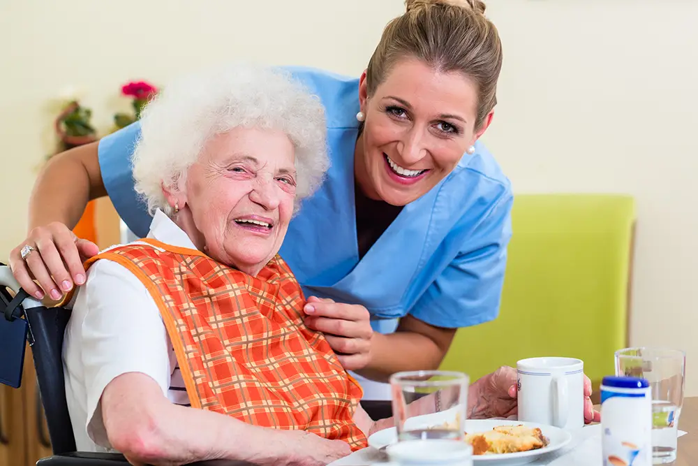 An elderly woman in a wheelchair smiles with a caregiver beside her. The woman wears an orange checked bib and sits at a table with food and drinks. An elderly woman in a wheelchair smiles with a caregiver beside her. The woman wears an orange checked bib and sits at a table with food and drinks.