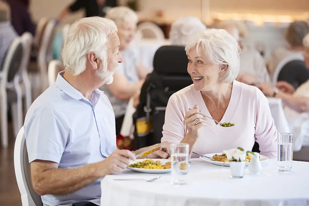 Two older adults sit at a table in a dining area, smiling and eating a meal together, with other people in the background.