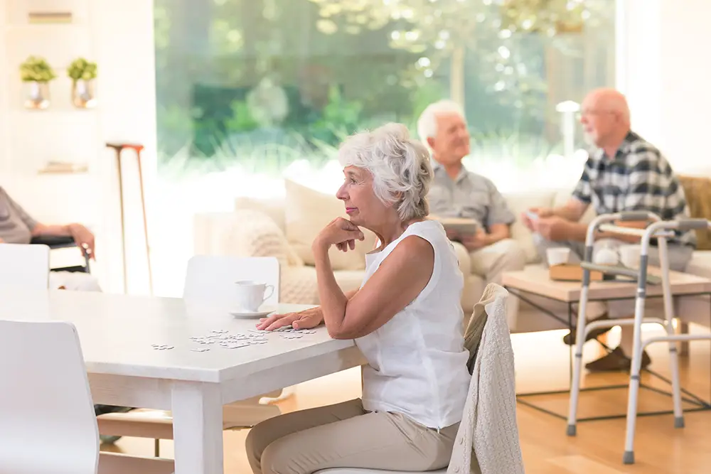 An older woman sits at a table working on a puzzle in a bright room, while three other older adults converse in the background. An older woman sits at a table working on a puzzle in a bright room, while three other older adults converse in the background.