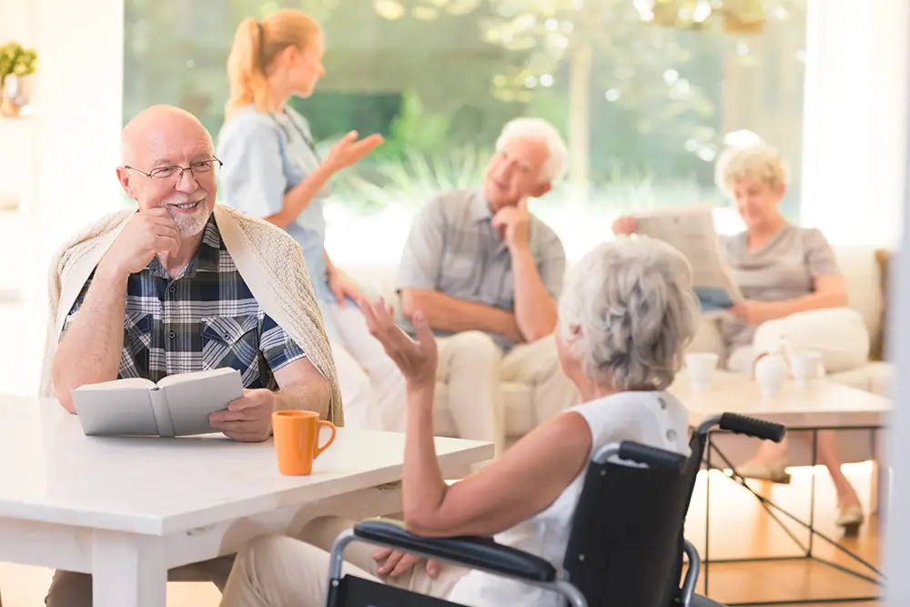 A group of older adults sit and converse in a bright common room; one man reads a book at a table with a woman in a wheelchair, while others talk and relax in the background.