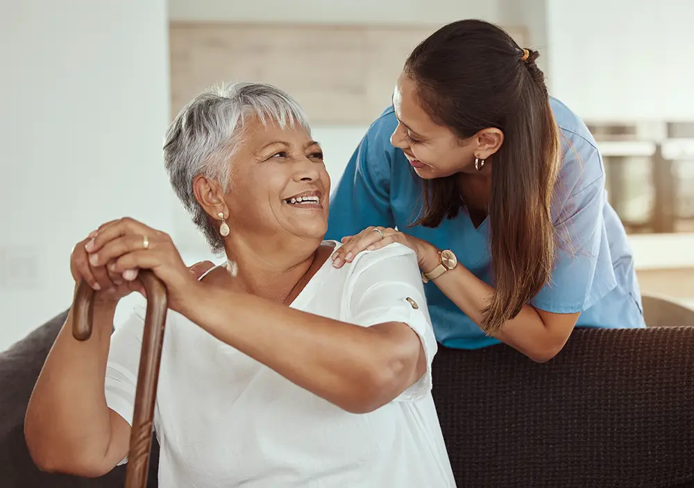 An older woman holding a cane sits on a couch while a caregiver in scrubs stands behind her, smiling and placing a hand on her shoulder. An older woman holding a cane sits on a couch while a caregiver in scrubs stands behind her, smiling and placing a hand on her shoulder.