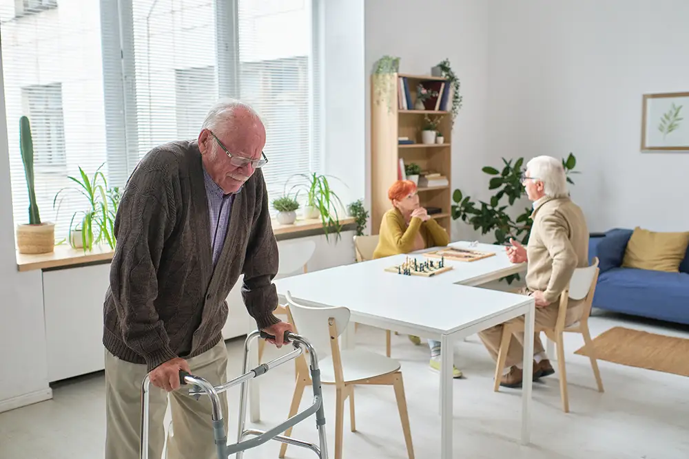 An elderly man uses a walker while two other seniors sit at a table playing chess in a bright, modern room with plants and large windows.