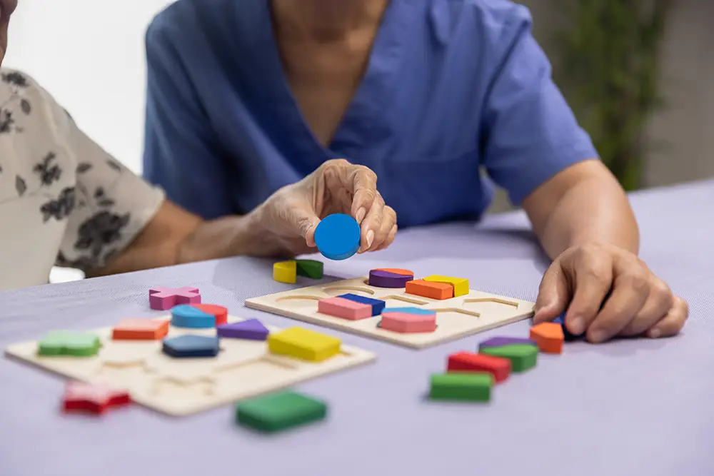 Two people sort colorful wooden shapes onto puzzle boards on a table. One person wears a blue uniform, the other has a floral-patterned top.