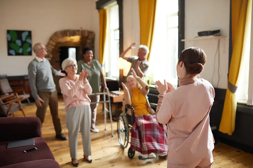 A group of seniors, including one in a wheelchair, clap and raise their arms while participating in an activity led by a caregiver in a sunny Arden Courts Memory Care room. A group of seniors, including one in a wheelchair, clap and raise their arms while participating in an activity led by a caregiver in a sunny Arden Courts Memory Care room.
