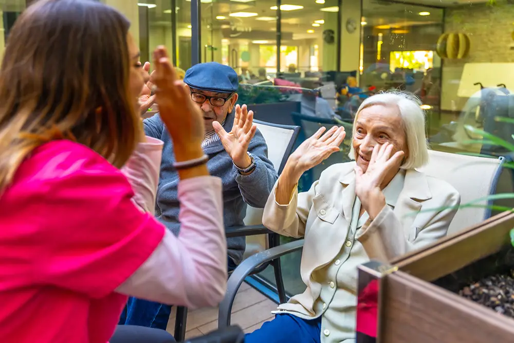 A caregiver and two elderly people sit outside, raising their hands and smiling during an interactive activity at a care facility.