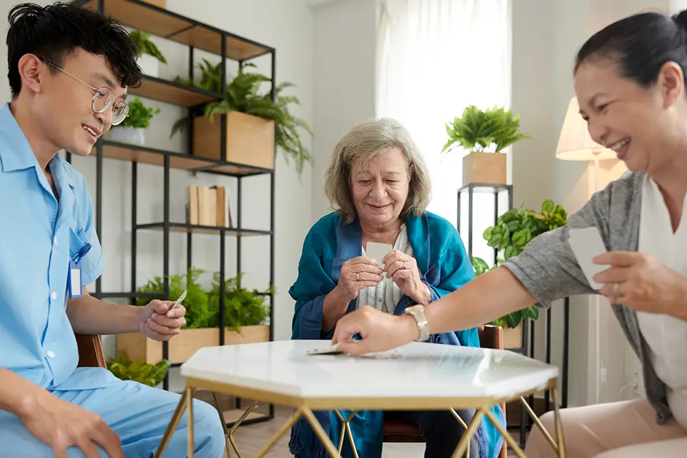 Three people sit around a table playing cards in a bright room with plants; one wears scrubs, another an elderly woman, and the third person is reaching to play a card. Three people sit around a table playing cards in a bright room with plants; one wears scrubs, another an elderly woman, and the third person is reaching to play a card.