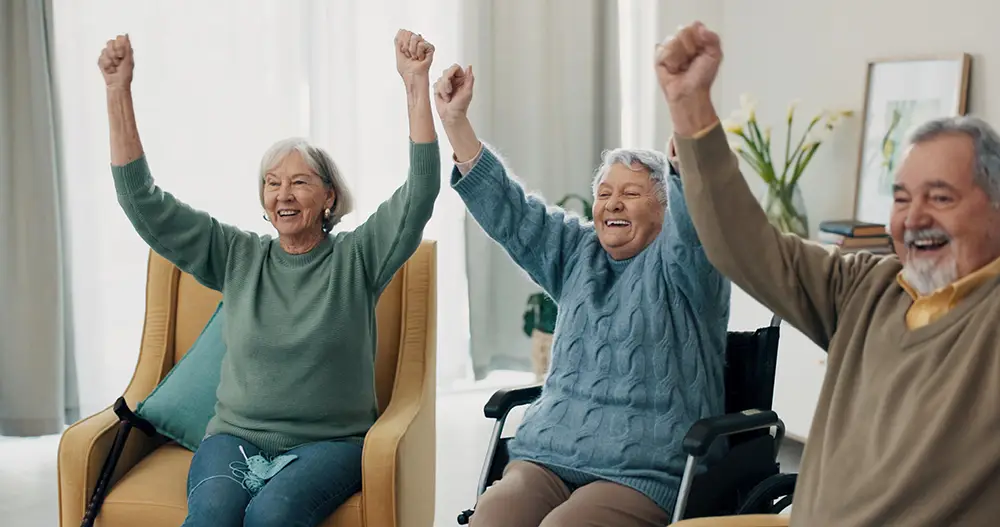 Three older adults, two women and one man, sit indoors with arms raised and smiling expressions, appearing engaged in a group activity.