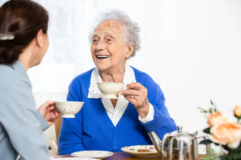 An older woman in a blue cardigan smiles while holding a teacup, sitting at a table with another person and a teapot.