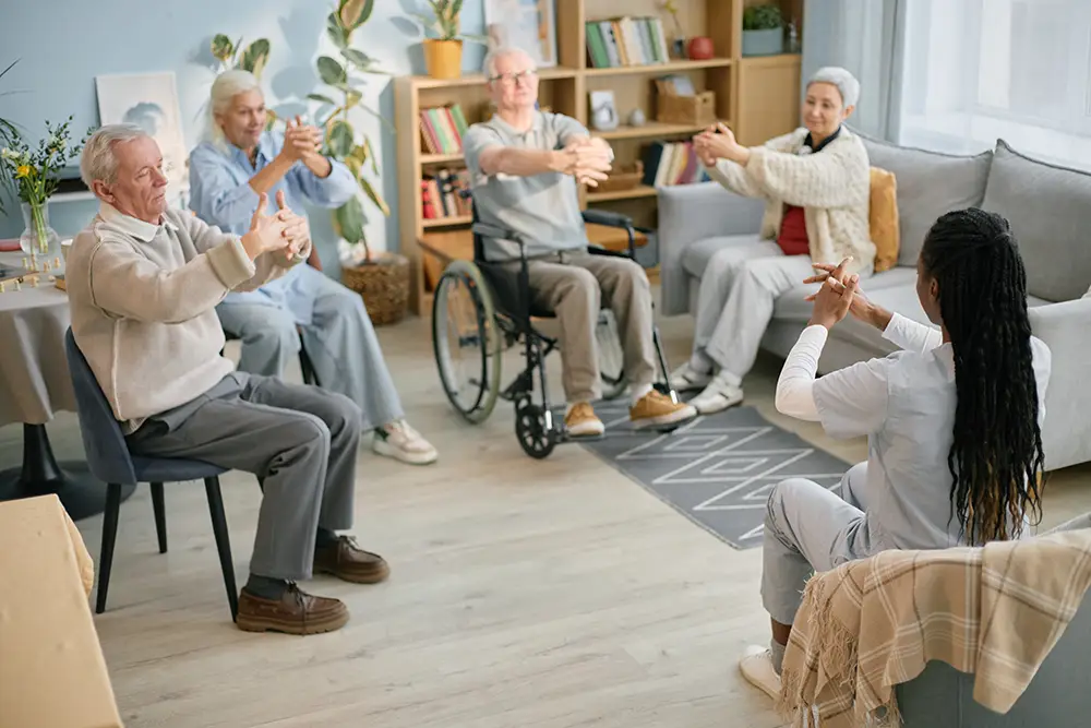 A group of seniors sit in a bright room, following a seated exercise led by an instructor at the front. One participant uses a wheelchair. Everyone is stretching their arms. A group of seniors sit in a bright room, following a seated exercise led by an instructor at the front. One participant uses a wheelchair. Everyone is stretching their arms.