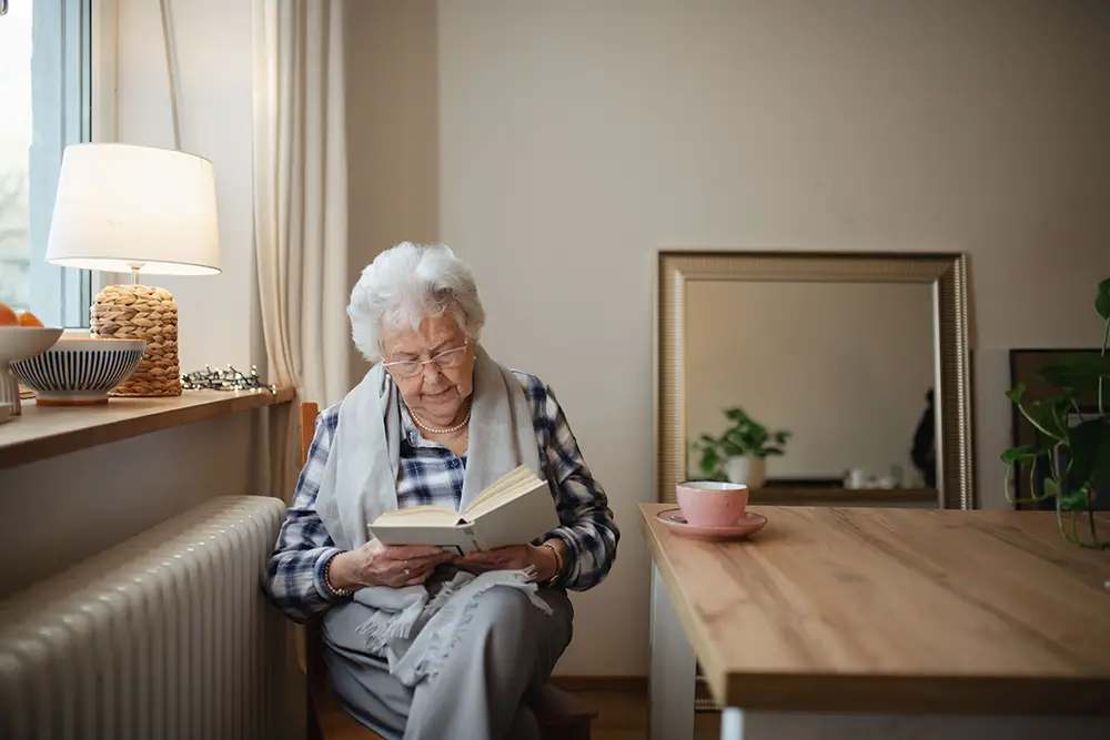 An elderly woman sits by a window reading a book, with a lamp, bowl, and cup on a wooden table nearby in a cozy, well-lit room.