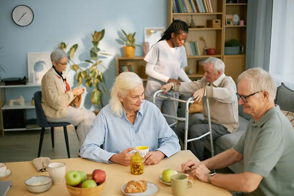 A group of elderly people and a caregiver interact in a well-lit communal room, with some seated at a table and others engaged in different activities.