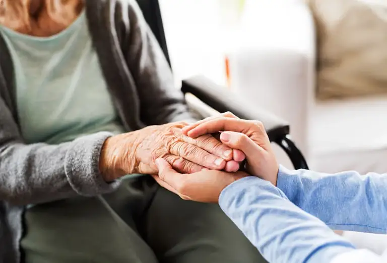 Close-up of a younger person gently holding the hands of an older adult sitting in a wheelchair, conveying support and care.