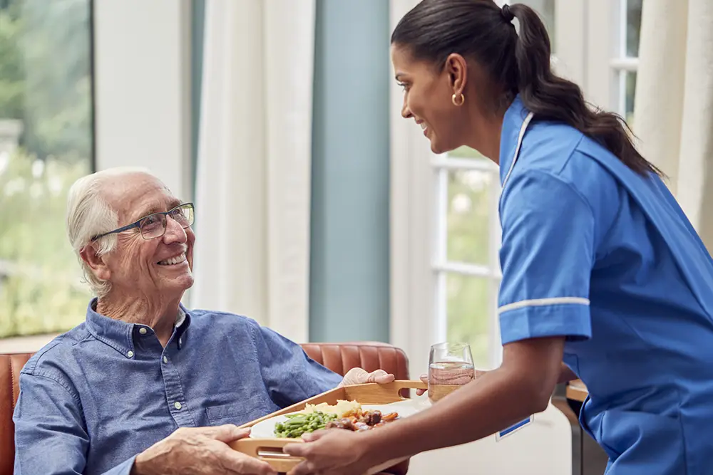 A caregiver in blue scrubs hands a meal tray and a glass of water to an elderly man sitting and smiling indoors.