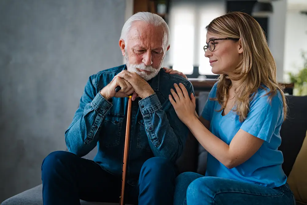 An elderly man sits with a cane, looking down thoughtfully, while a younger woman sits beside him, resting her hand supportively on his shoulder.
