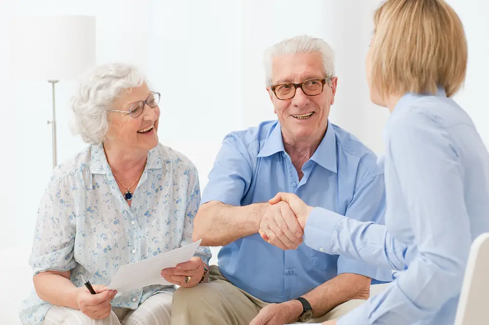 An elderly couple sits and smiles as the man shakes hands with a woman in business attire, while the woman holds paperwork. An elderly couple sits and smiles as the man shakes hands with a woman in business attire, while the woman holds paperwork.