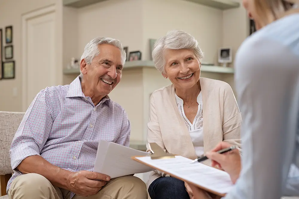 Two older adults sit on a couch, smiling, holding documents, while a person with a clipboard sits across from them taking notes. Two older adults sit on a couch, smiling, holding documents, while a person with a clipboard sits across from them taking notes.