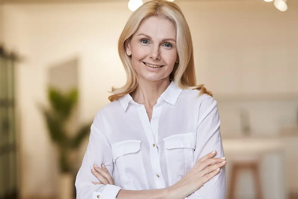 A woman with blonde hair wearing a white button-up shirt stands indoors with her arms crossed, smiling at the camera.