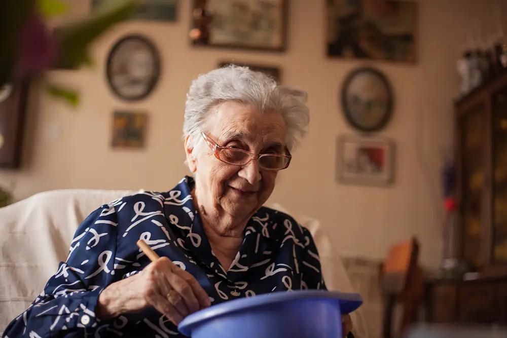 An elderly woman with gray hair and glasses sits indoors, smiling while mixing something in a blue bowl with a spoon.