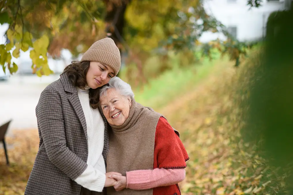 An elderly woman smiles and hugs a younger woman outdoors among autumn trees and fallen leaves. An elderly woman smiles and hugs a younger woman outdoors among autumn trees and fallen leaves.