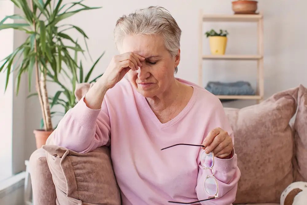 Older woman sitting on a couch, holding her glasses in one hand and pinching the bridge of her nose with her eyes closed, appearing tired or stressed. Older woman sitting on a couch, holding her glasses in one hand and pinching the bridge of her nose with her eyes closed, appearing tired or stressed.