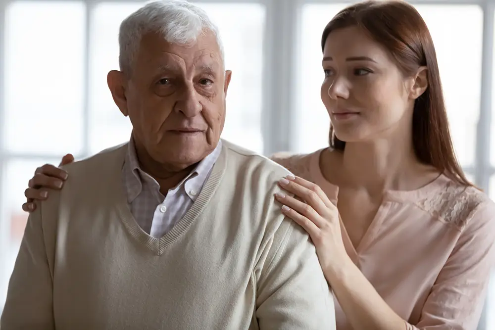 An older man with gray hair sits while a younger woman with long brown hair stands behind him, resting her hands on his shoulders. Both have neutral expressions. An older man with gray hair sits while a younger woman with long brown hair stands behind him, resting her hands on his shoulders. Both have neutral expressions.
