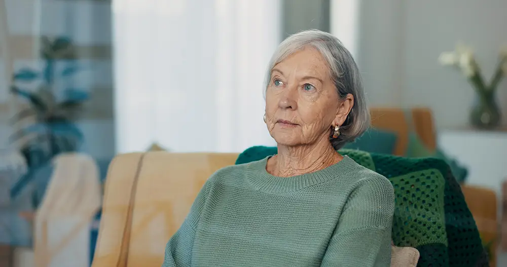 An older woman with gray hair and a green sweater sits indoors on a couch, looking slightly upward with a neutral expression. An older woman with gray hair and a green sweater sits indoors on a couch, looking slightly upward with a neutral expression.
