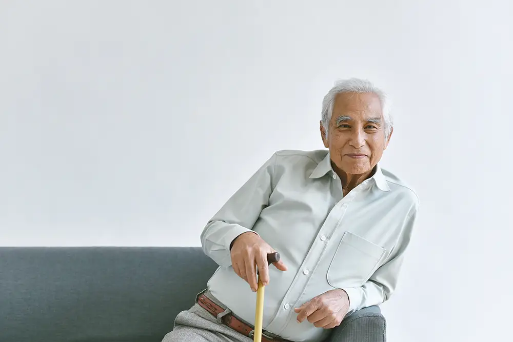 Older man with gray hair, wearing a light shirt, sits on a gray sofa and holds a cane, looking at the camera against a plain white background. Older man with gray hair, wearing a light shirt, sits on a gray sofa and holds a cane, looking at the camera against a plain white background.