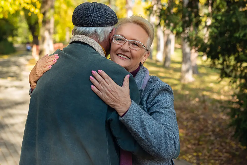 An older woman hugs an older man outdoors on a sunny day, smiling at the camera. Trees and a path are visible in the background. An older woman hugs an older man outdoors on a sunny day, smiling at the camera. Trees and a path are visible in the background.