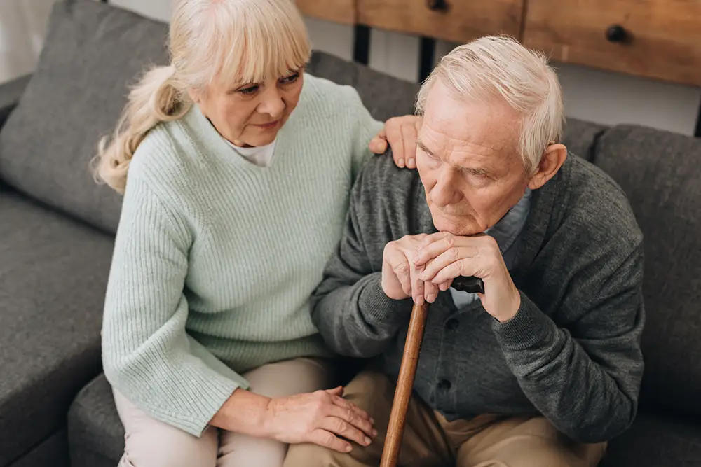An elderly man sits on a couch holding a cane, looking down, while an elderly woman sits beside him with a supportive hand on his shoulder and knee. An elderly man sits on a couch holding a cane, looking down, while an elderly woman sits beside him with a supportive hand on his shoulder and knee.
