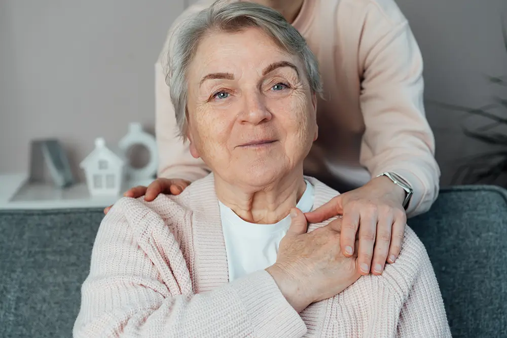 An older woman sits on a couch, smiling gently, with another person’s hands resting on her shoulders in a supportive gesture. An older woman sits on a couch, smiling gently, with another person's hands resting on her shoulders in a supportive gesture.