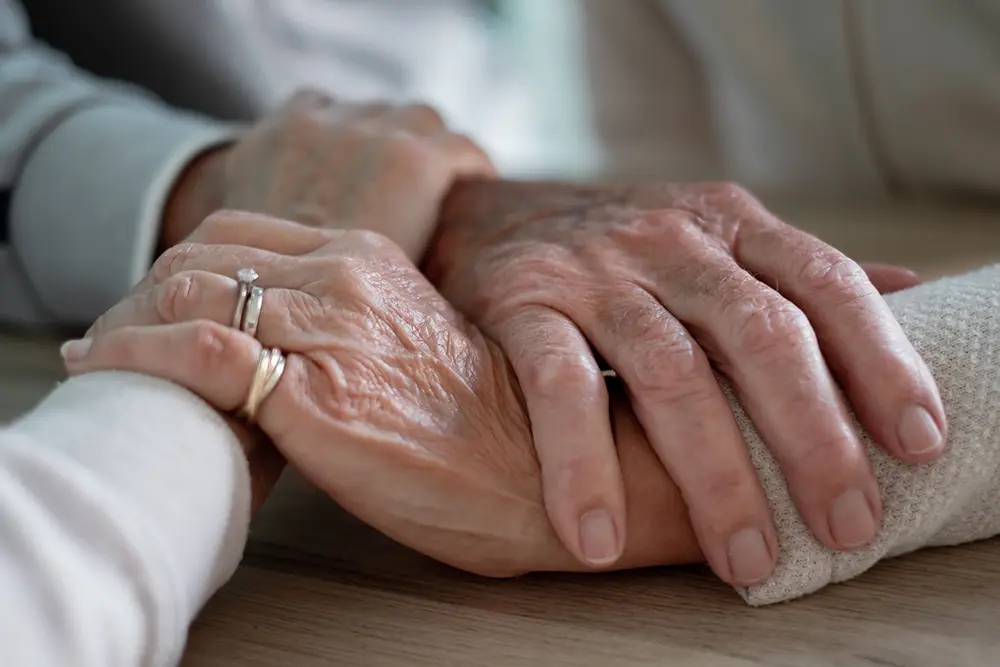 Close-up of two elderly people holding hands on a wooden surface, showing support and connection. Close-up of two elderly people holding hands on a wooden surface, showing support and connection.