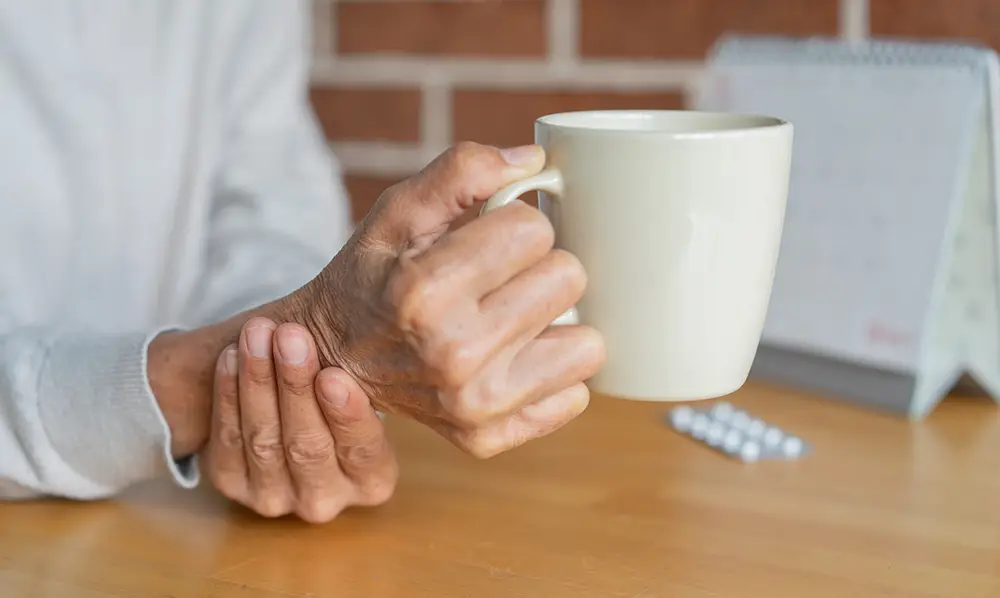 A person holds a white mug in one hand while gripping their wrist with the other; pills and a calendar are on the wooden table in the background. A person holds a white mug in one hand while gripping their wrist with the other; pills and a calendar are on the wooden table in the background.