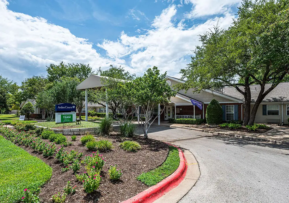 Single-story building with a covered entrance, surrounded by trees, landscaped flower beds, and a sign that reads “Arden Courts.” The sky is partly cloudy.