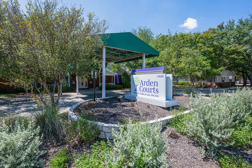 The entrance of Arden Courts, a memory care community in Austin, features a green canopy and a sign reading “Arden Courts,” surrounded by lush landscaping and trees under a blue sky.