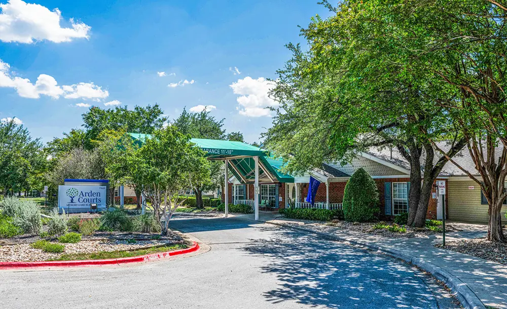 Entrance view of Arden Courts, a residential care facility, with a covered driveway, sign, trees, and landscaped surroundings under a clear blue sky.