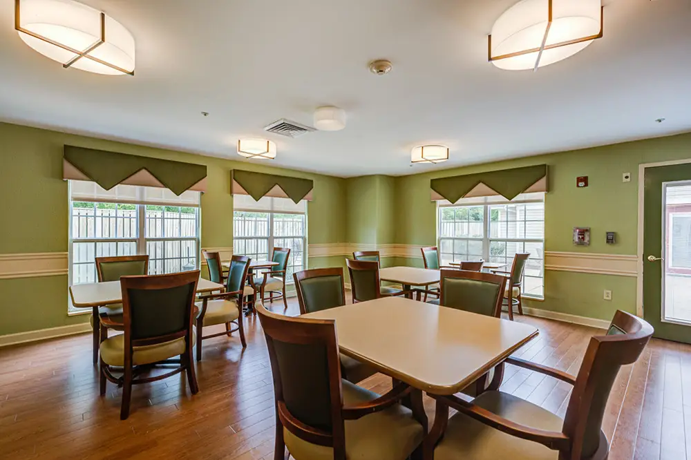 A dining room with several square tables and wooden chairs, green walls, three large windows with valances, and ceiling lights. The room has wood flooring and natural light.