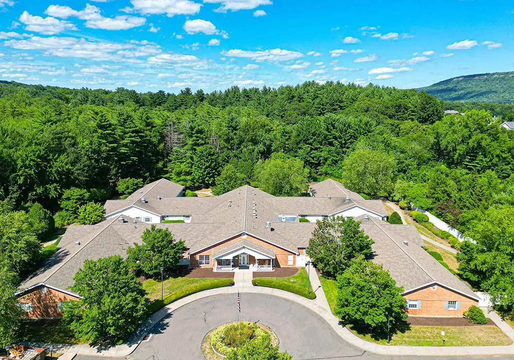 Aerial view of Arden Courts, a single-story brick memory care building in Avon, surrounded by trees and greenery, with a circular driveway in front and wooded hills in the background.