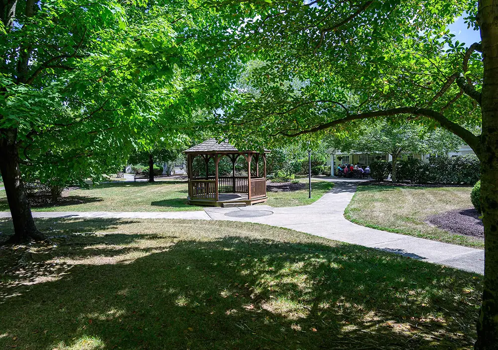 A wooden gazebo sits at the center of a grassy park area, surrounded by trees and intersected by concrete walkways on a sunny day. A wooden gazebo sits at the center of a grassy park area, surrounded by trees and intersected by concrete walkways on a sunny day.