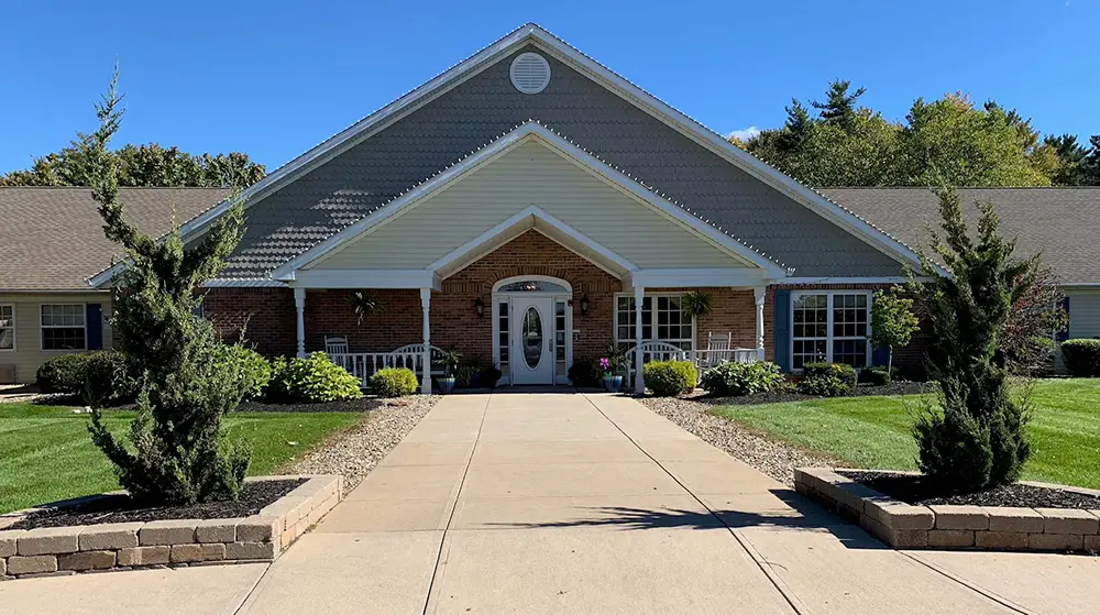 Single-story brick building with a peaked roof, landscaped entrance, and symmetrical pathways leading to double doors—an inviting day at Arden Courts, designed for memory care residents in Bath.