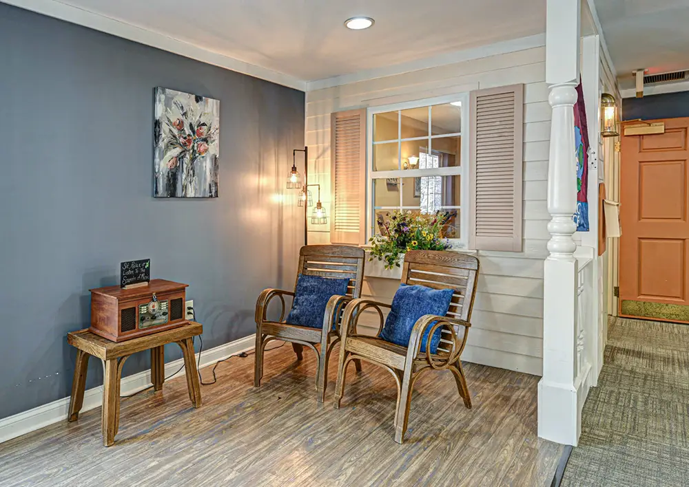 A small seating area with two wooden chairs, blue cushions, a side table, a vintage radio, wall art, flowers, and decorative lighting against gray and beige walls.