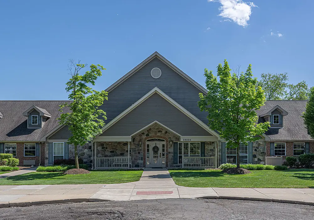 A single-story building with stone and gray siding, a pitched roof, front porch, and two trees in front under a clear blue sky—perfect for memory care at Arden Courts in Bingham Farms.