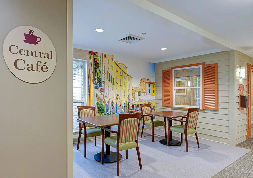 Small café seating area with four wooden tables and green chairs, mural of colorful buildings on wall, and “Central Café” sign in foreground. Small café seating area with four wooden tables and green chairs, mural of colorful buildings on wall, and "Central Café" sign in foreground.