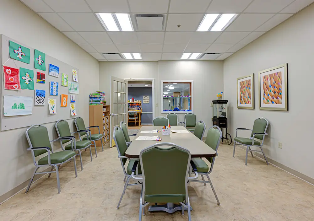 A classroom with green chairs around a table, children’s artwork on the walls, and craft supplies on the table under fluorescent ceiling lights.