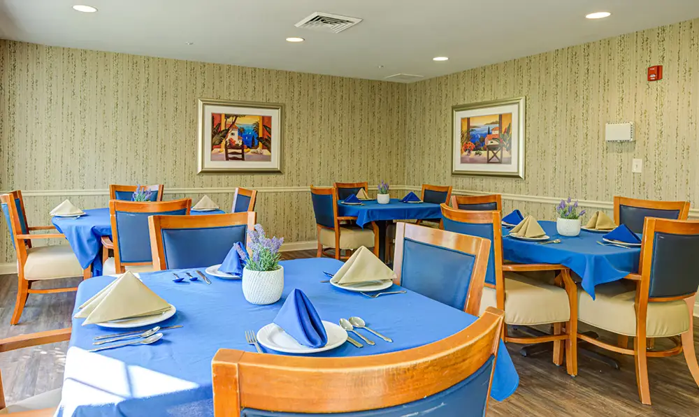 Dining room with several tables covered in blue tablecloths, each set with plates, utensils, and napkins, and decorated with small potted plants; framed art on the walls.